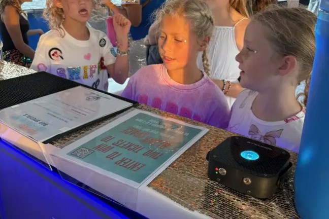 Group of children standing at a bar on a boat with waterfront buildings in the background.