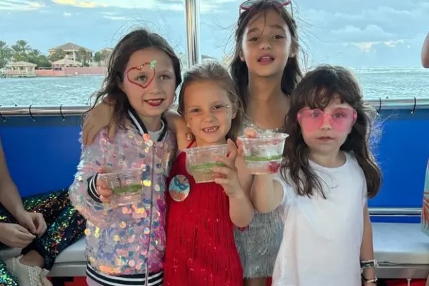 Four children on a boat, dressed festively, holding drinks, with a water view in the background.