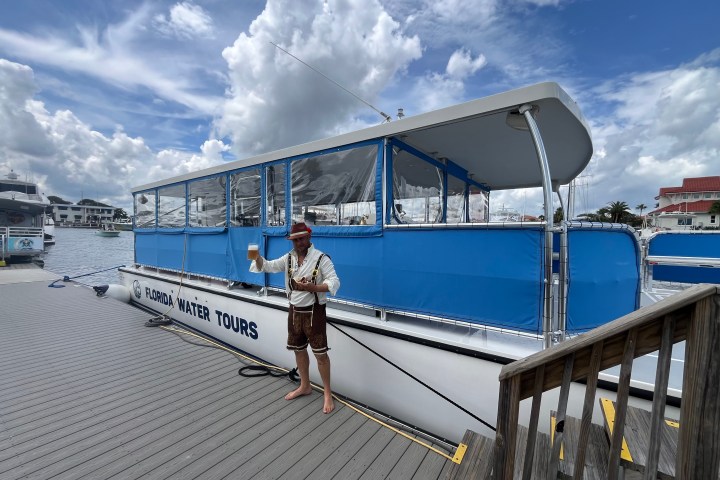 Person in traditional attire holding a drink next to a tour boat docked at a marina.