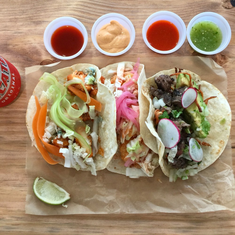 Three tacos with various toppings, sauces in cups, and a lime wedge on a wooden table.