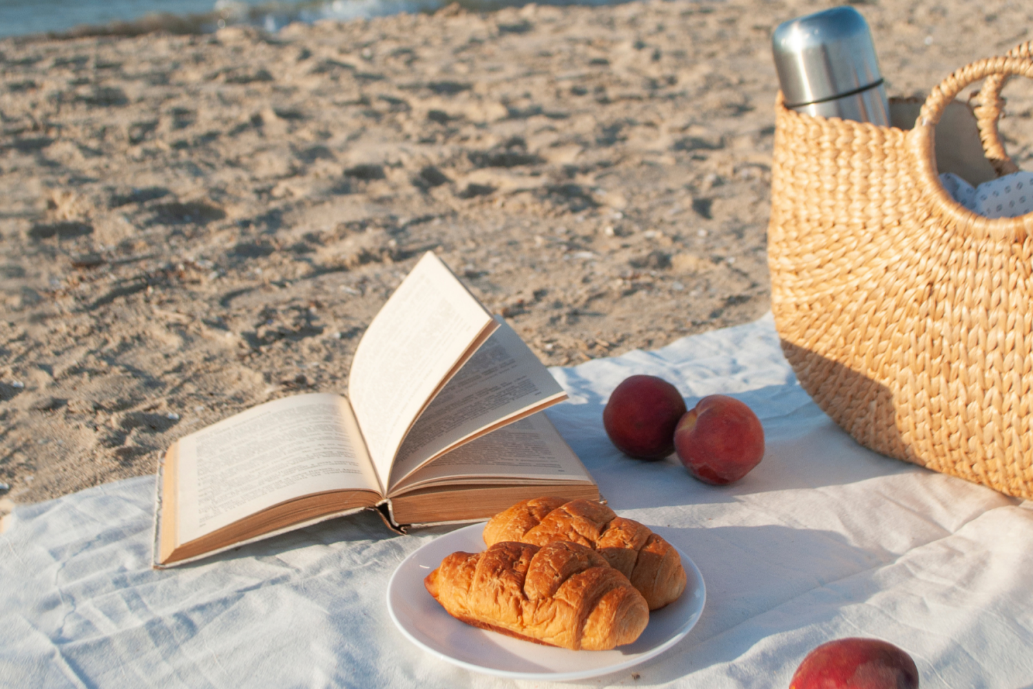 Book, croissants, and basket on beach blanket near the sea.
