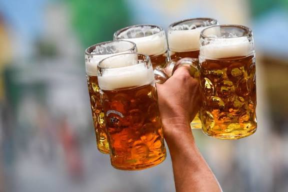 Hand holding four large beer mugs with foamy tops in an outdoor setting.