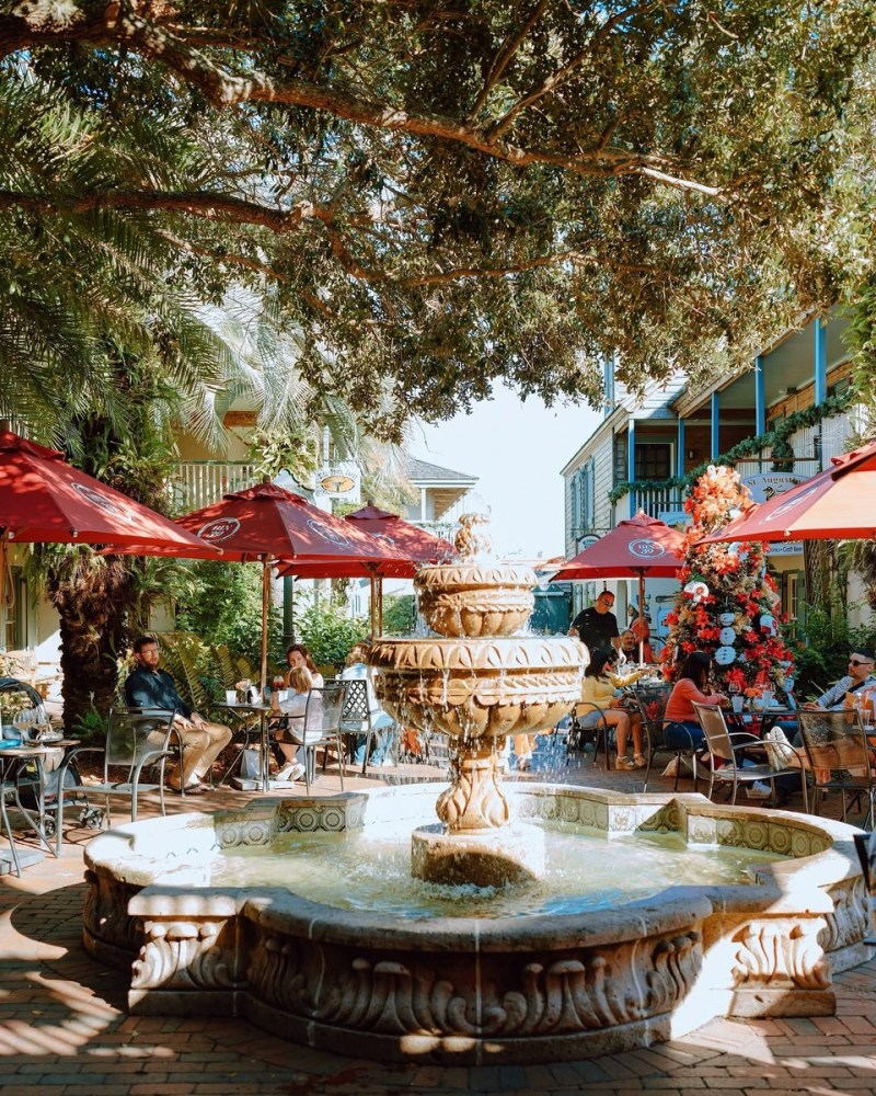 Outdoor cafe with a stone fountain, red umbrellas, and lush greenery.