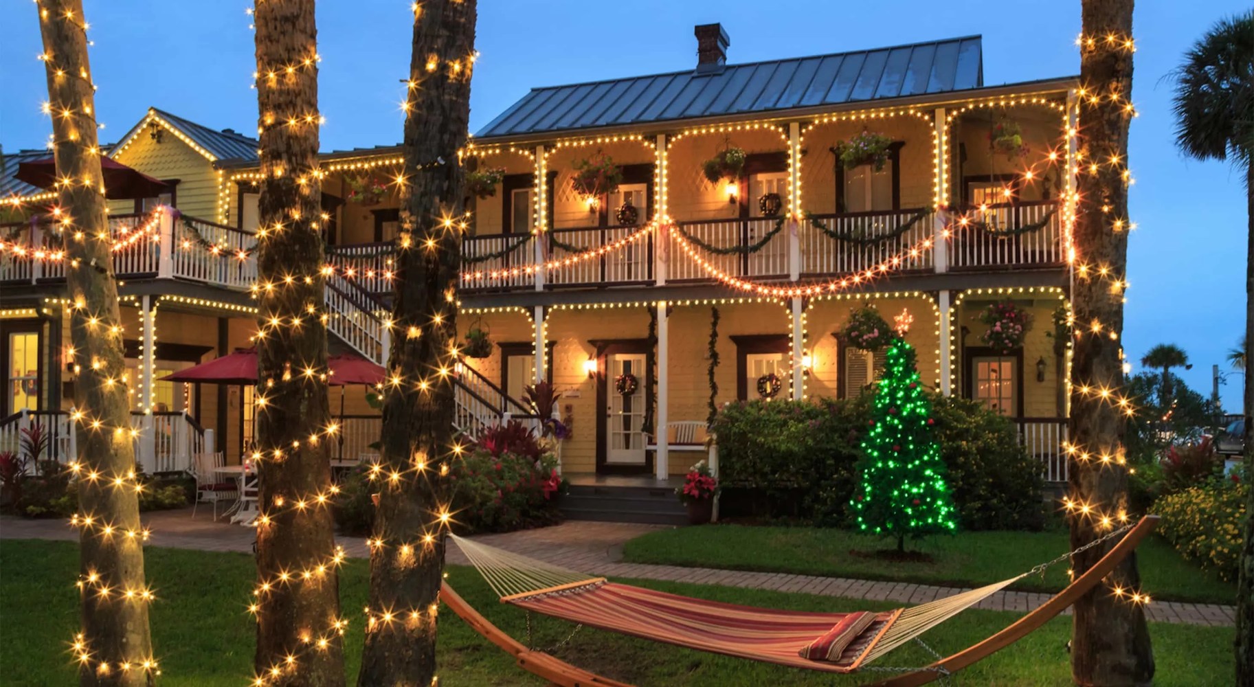 House with festive lights and palm trees, hammock on lawn.